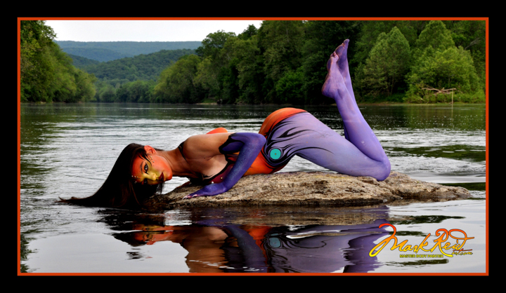 Dark haried woman on a rock that is barely out of the water in a river body painted in purple and red with her hair in the water.